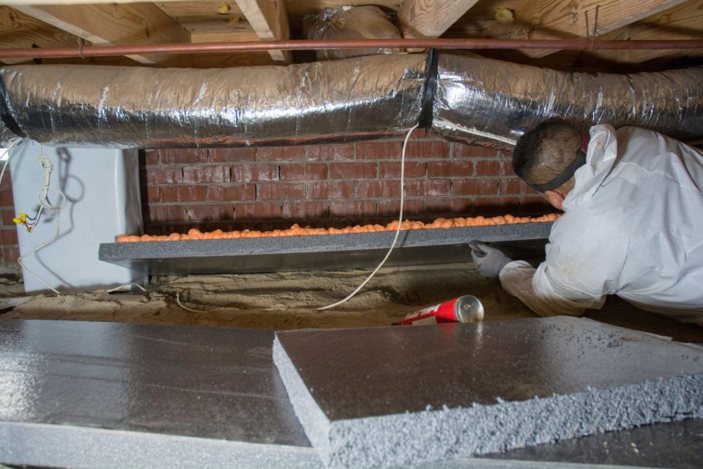 A worker in protective gear installs insulation beneath a floor, surrounded by ductwork and foam boards in a dimly lit basement.