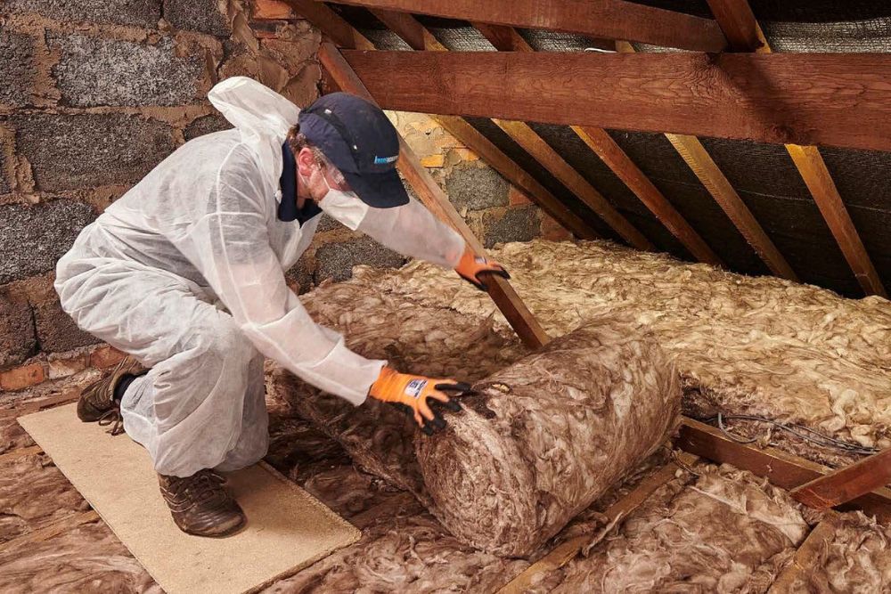 A man in a white coat and safety glasses installs insulation in an attic, focusing on safety and precision in his work.