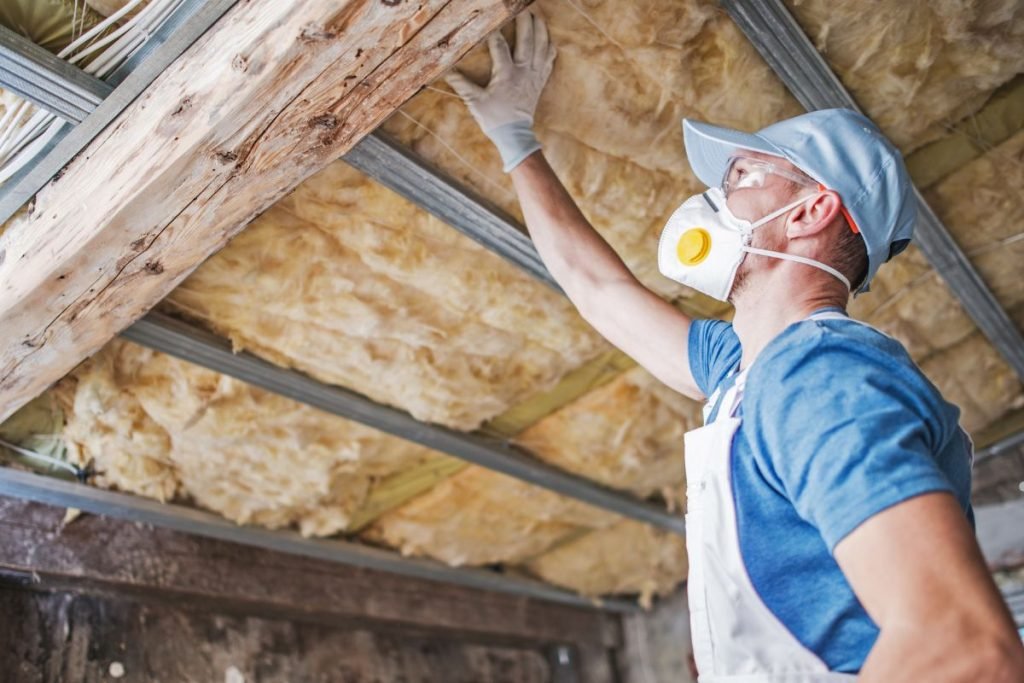 A worker wearing a mask and gloves installs insulation in a ceiling, surrounded by soft yellow insulation material.